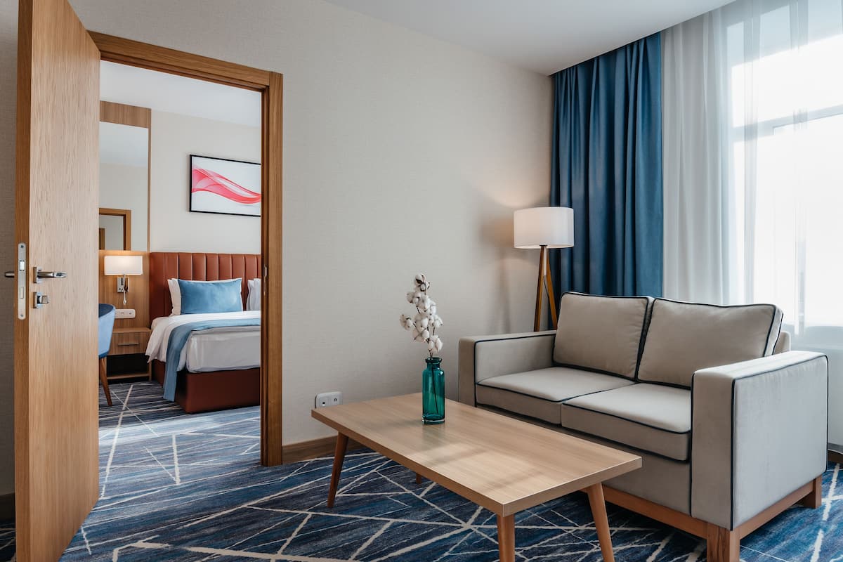 Une chambre d’hôtel avec un lit, un canapé, une table basse, un vase avec des fleurs, une lampe et une fenêtre avec des rideaux.