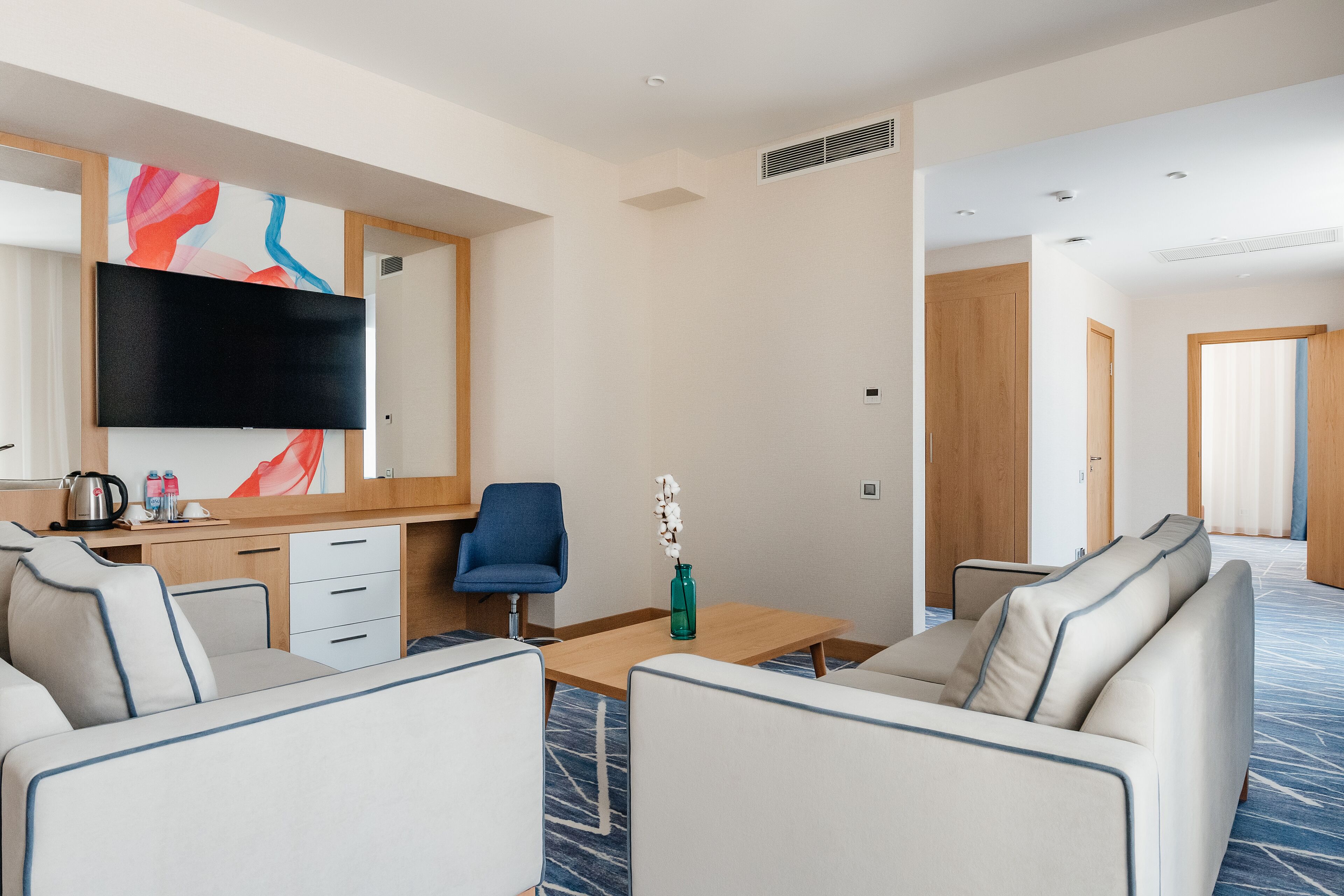 Une chambre d’hôtel moderne équipée d’un téléviseur à écran plat, d’un bureau en bois, d’une chaise bleue, d’un canapé et d’un vase décoratif.