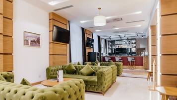A modern hotel lobby with green upholstered sofas, a flat-screen TV, and a bar area in the background.