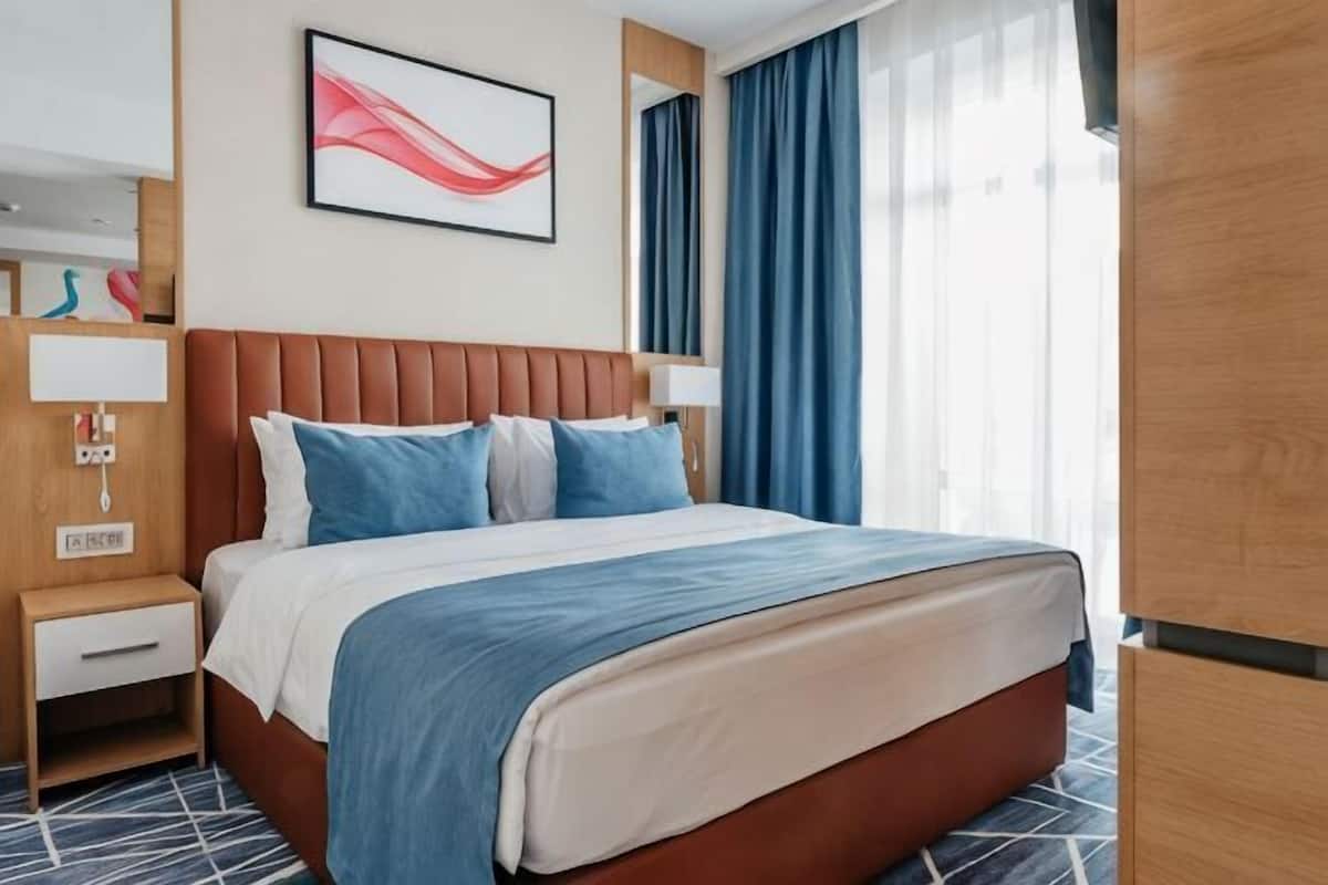Une chambre d’hôtel avec un lit, une table de chevet, une lampe, un téléviseur fixé au mur et une fenêtre avec des rideaux.