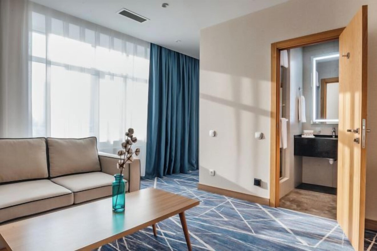 A modern hotel room with a sofa, a wooden table, a vase with flowers, a large window with blue curtains, and a bathroom visible through an open door.