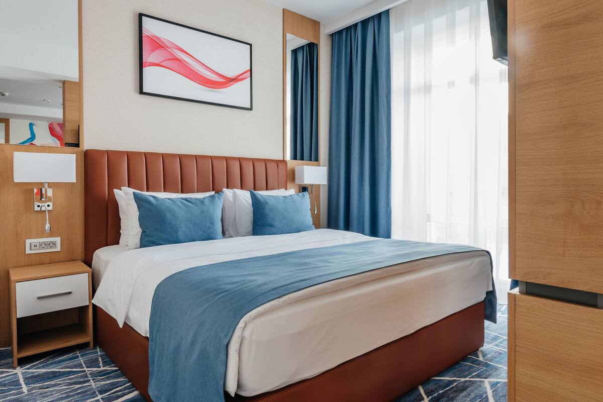 Une chambre d’hôtel avec un grand lit, une table de chevet avec une lampe, un miroir fixé au mur et une fenêtre avec des rideaux.
