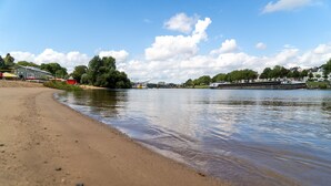 Ein Flussstrand mit einem Boot, das im Wasser liegt, einem Sandufer und Gebäuden im Hintergrund.