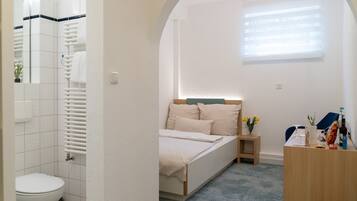 A bedroom with a bed, a bedside table with a lamp, a window with blinds, and a bathroom with a toilet and towel rack.