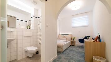 Uma casa de banho moderna com sanita, lavatório e chuveiro. Há uma cama com uma mesa de cabeceira e uma pequena mesa com uma lâmpada.