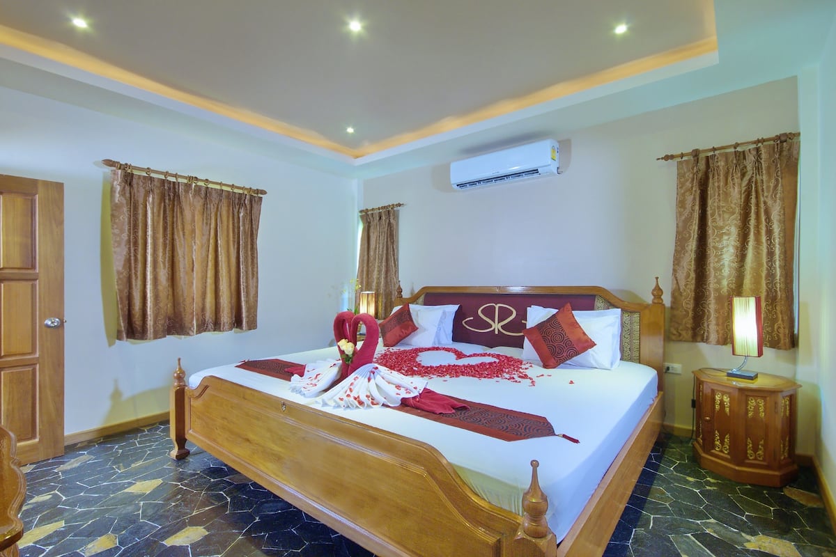 Quarto de hotel bem iluminado com uma cama grande, um guarda-roupa de madeira e uma mesa de cabeceira com abajur.