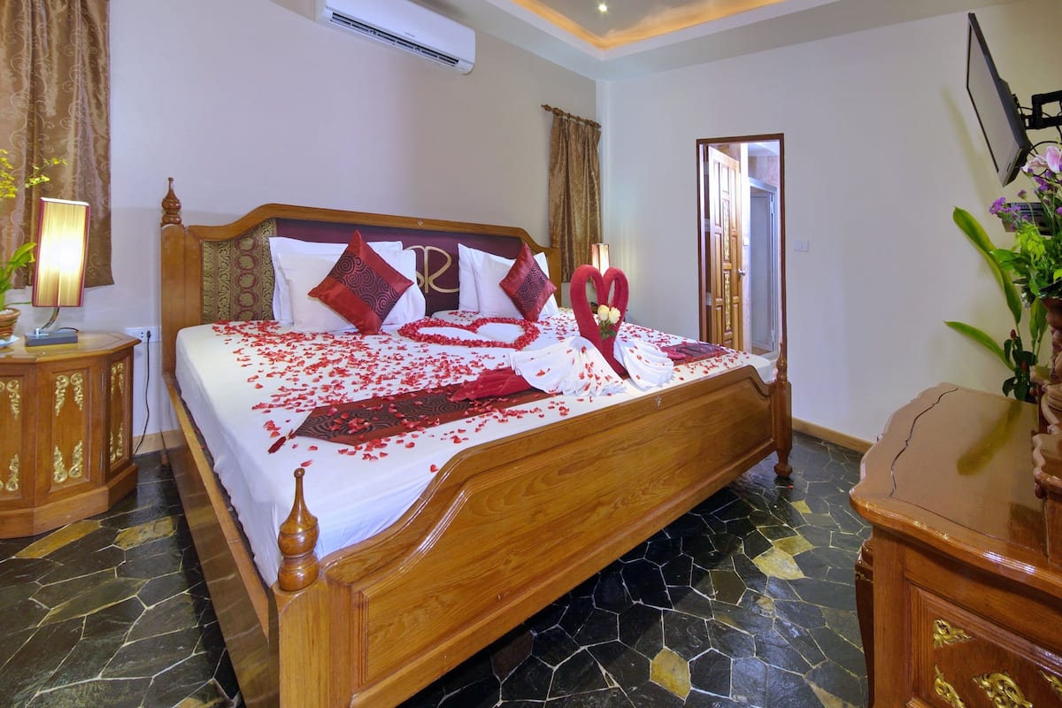 Uma cama bem arrumada com cabeceira de madeira, mesa de cabeceira com abajur e uma planta decorativa.