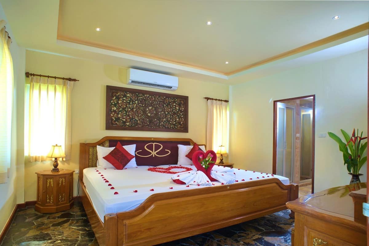 Quarto de hotel com cama, cabeceira de madeira, ar condicionado, um objeto decorativo na parede, espelho e um vaso com flores.