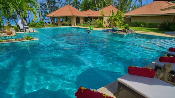 A clear swimming pool with lounge chairs and towels, surrounded by palm trees and a building with a red roof.
