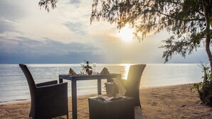 A beachside table with chairs, a vase with flowers, and a bottle, set against a sunset view.