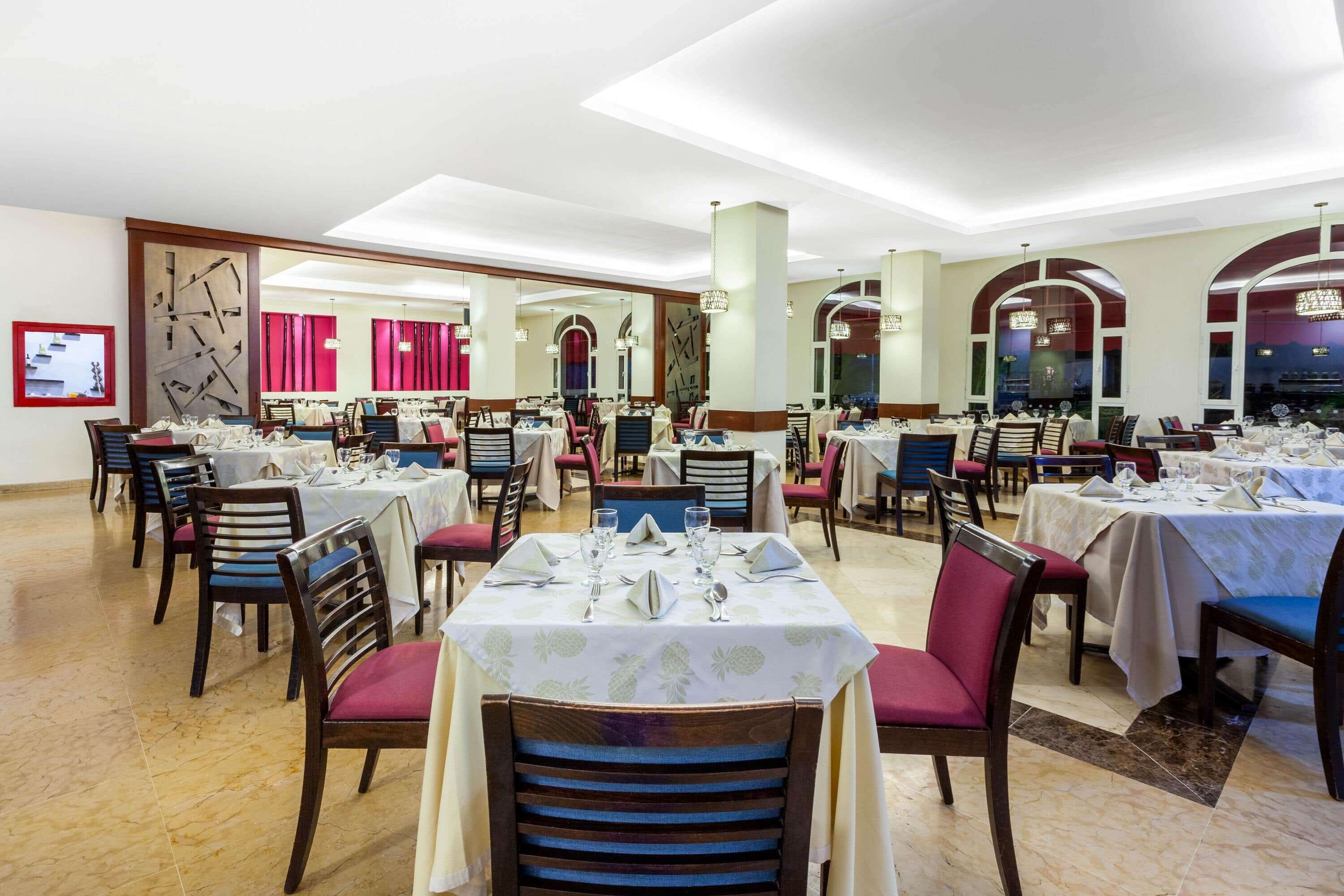 Un restaurante con varias mesas preparadas para comer, que cuenta con una decoración moderna con candelabros y ventanas arqueadas.