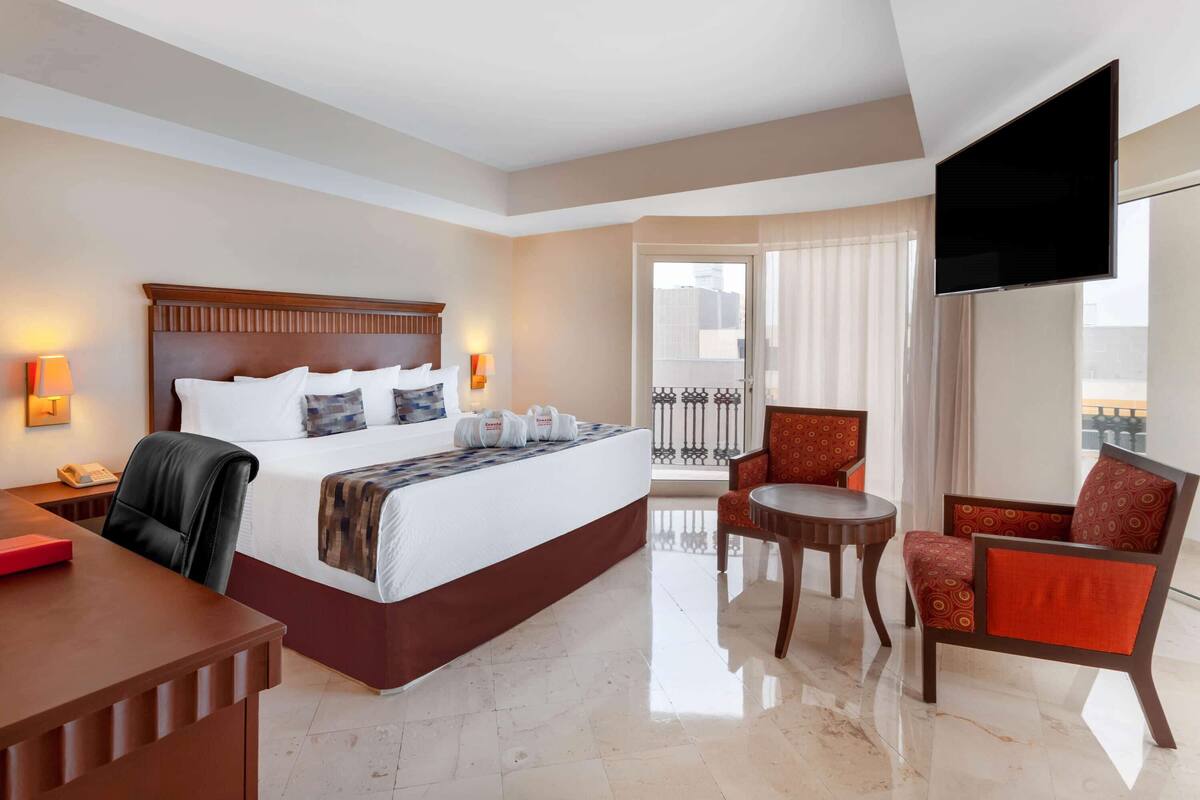 A hotel room with a bed, a desk, a chair, a TV, and a balcony with curtains.