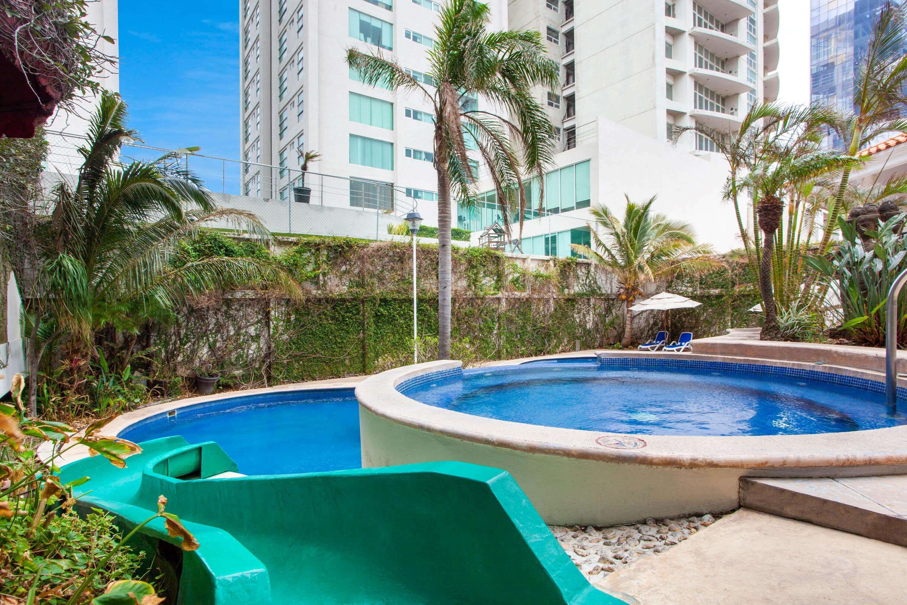 A swimming pool with a slide, surrounded by greenery and tall buildings.