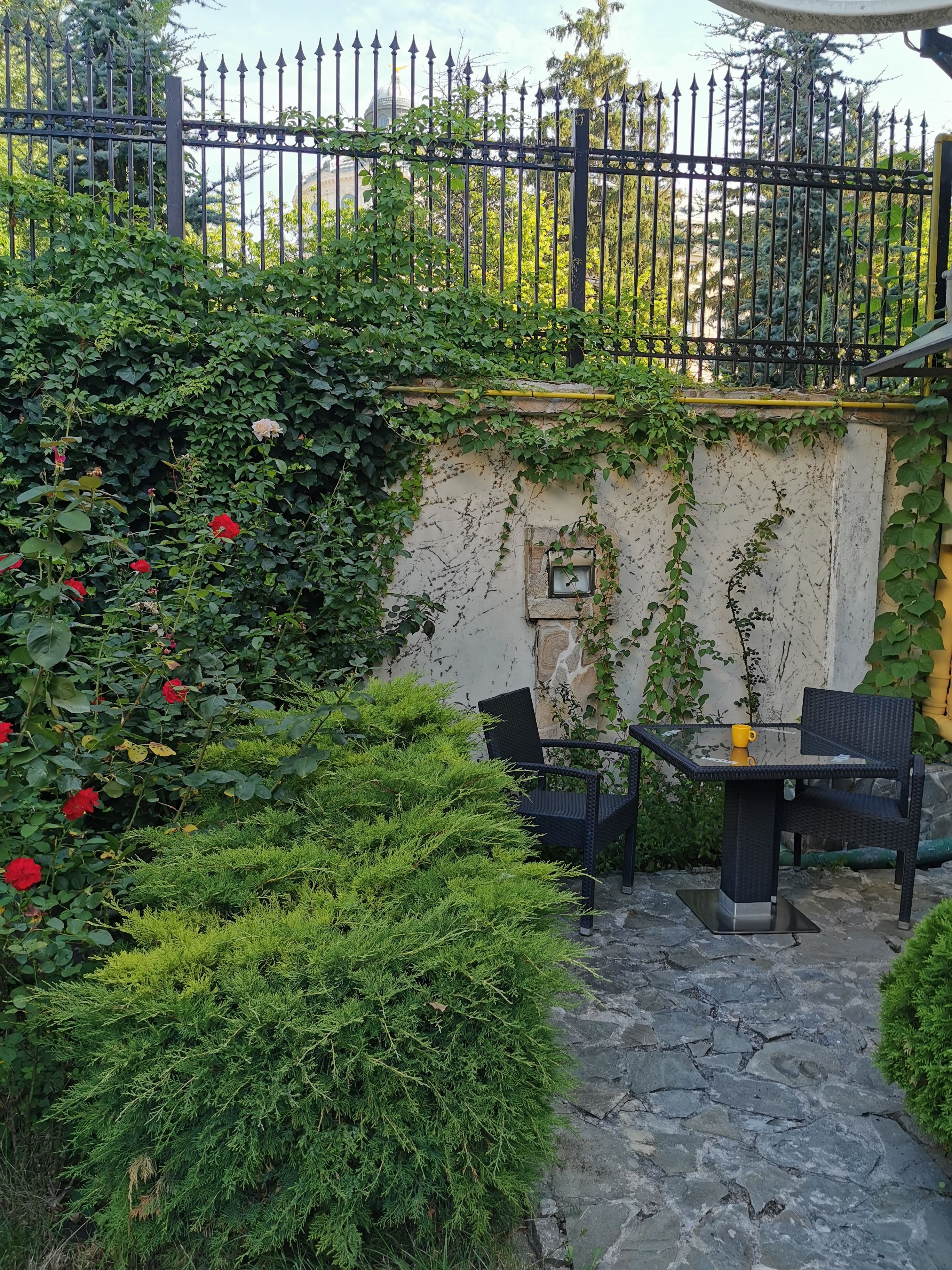 A stone patio with a table and chairs, surrounded by lush greenery and a black metal fence.