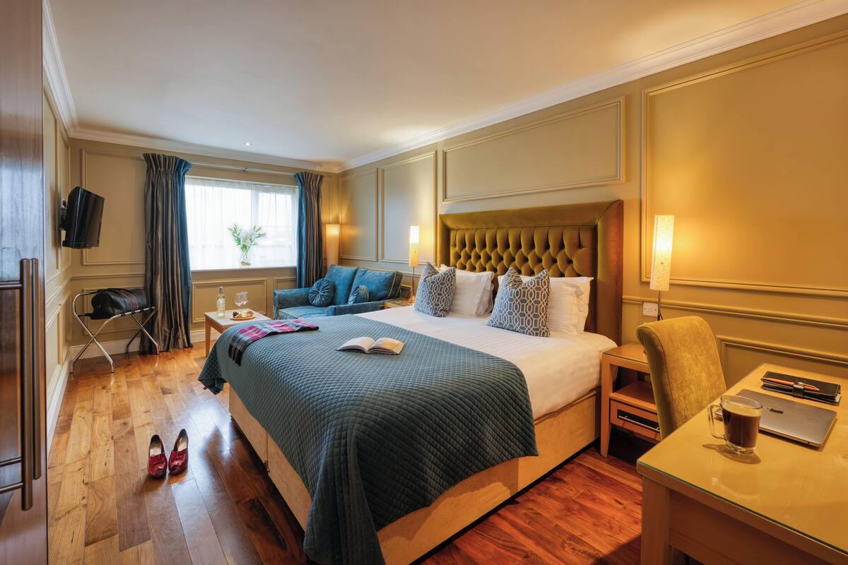 Una habitación de hotel con una cama grande, un escritorio con una computadora portátil, una silla, un televisor montado en la pared y una ventana con cortinas.