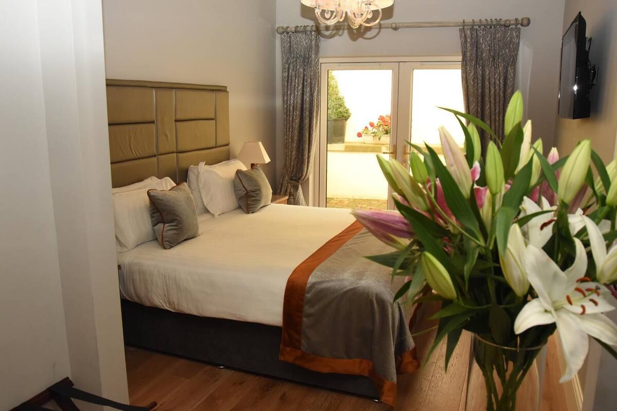 Un dormitorio con cama, mesita de noche, lámpara, ventana con cortinas y una planta en maceta.