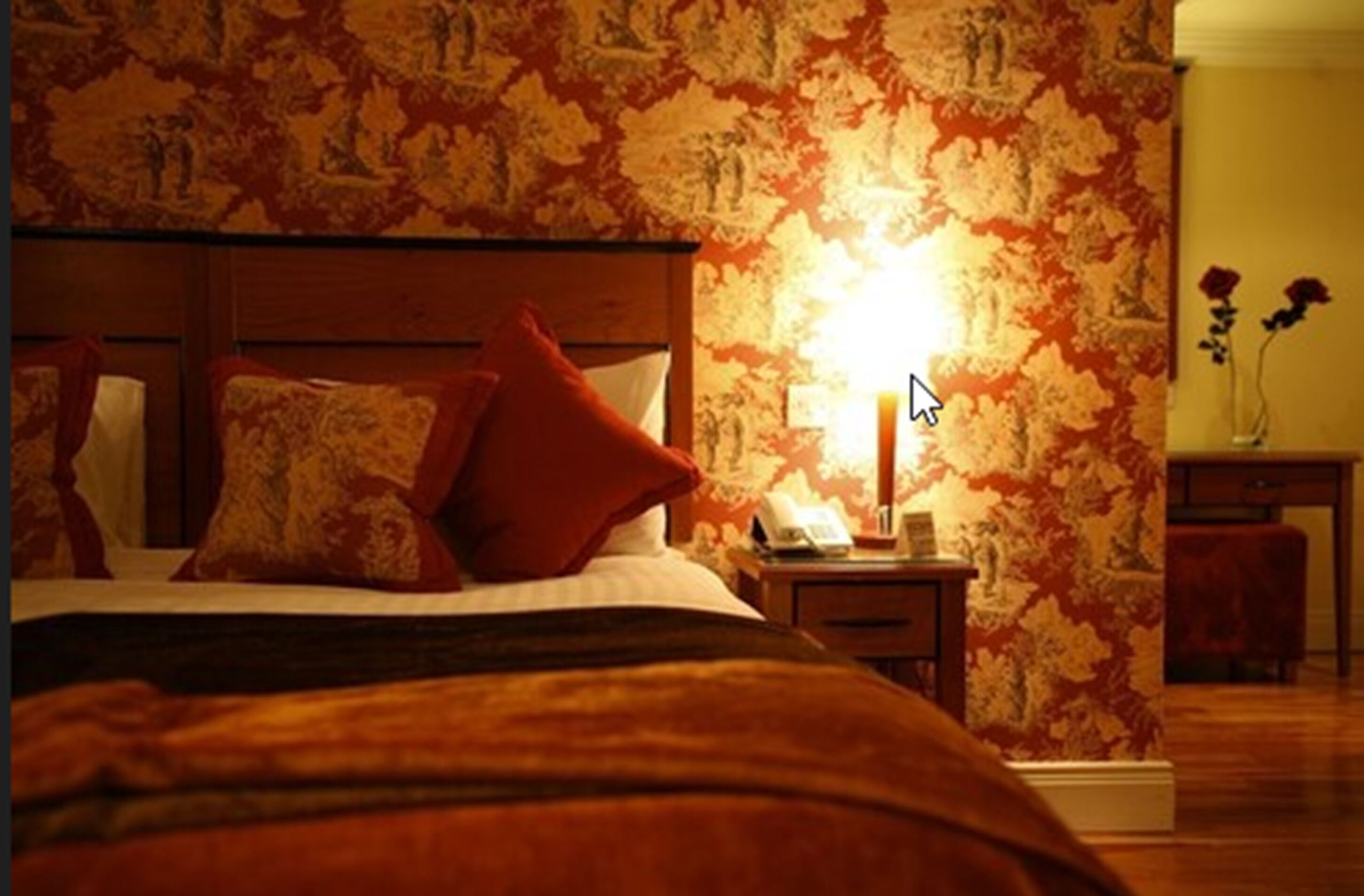 A bedroom with a bed, a nightstand, a lamp, and a floral wallpaper.