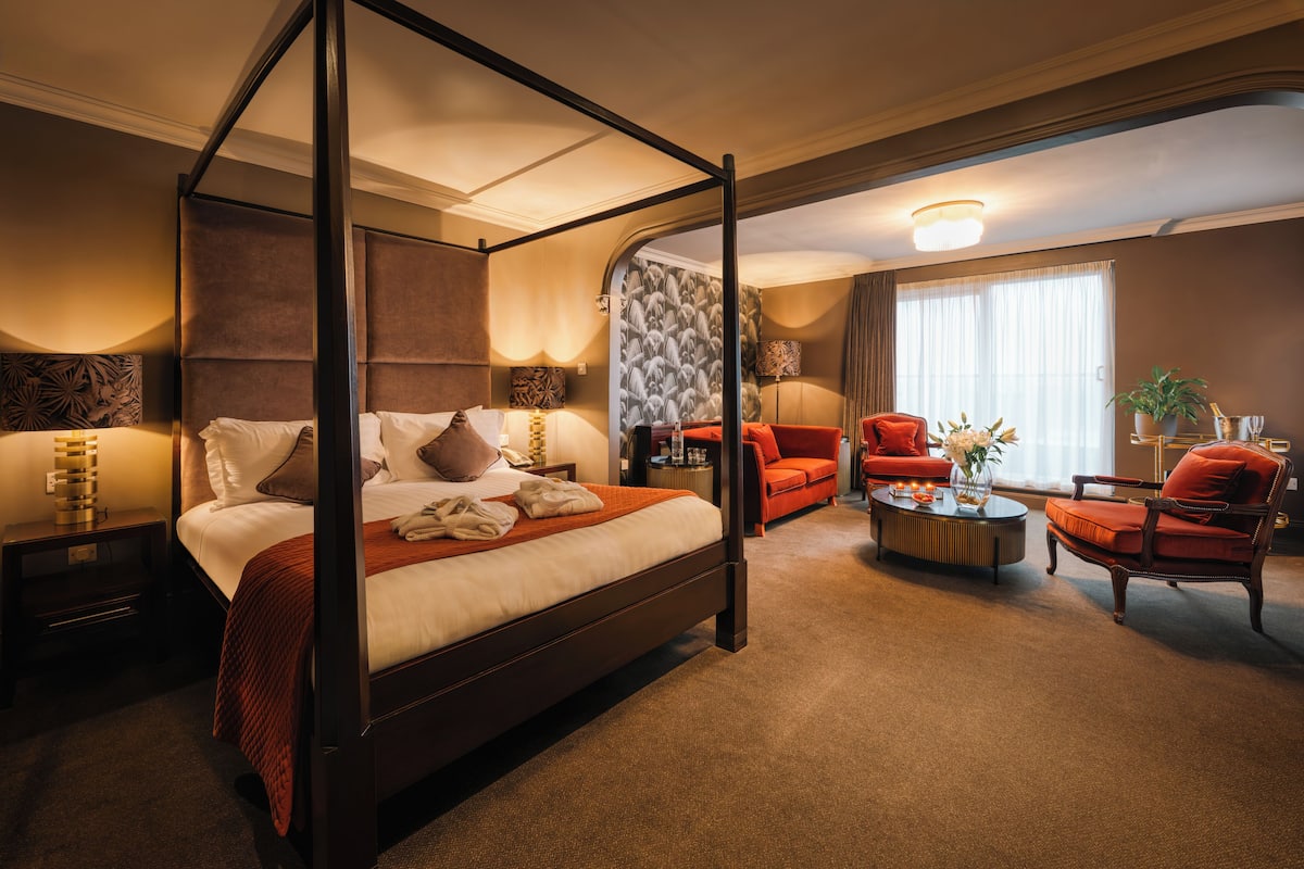 Una cama con dosel, una manta roja, una mesita de centro redonda y dos sillones rojos en una habitación de hotel.
