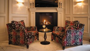 Una habitación con dos sillones estampados, una mesita redonda con un vaso y una vela, y una chimenea encendida.