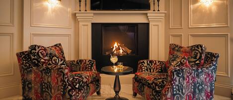 A room with two patterned armchairs, a small round table with a glass and a candle, and a fireplace with a fire.