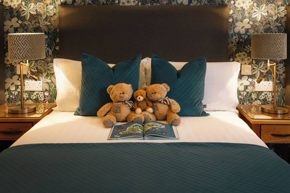 Una cama cuidadosamente hecha con un edredón azul acolchado, dos almohadas color turquesa y tres osos de peluche sentados sobre ella. Un libro abierto está colocado sobre la cama.