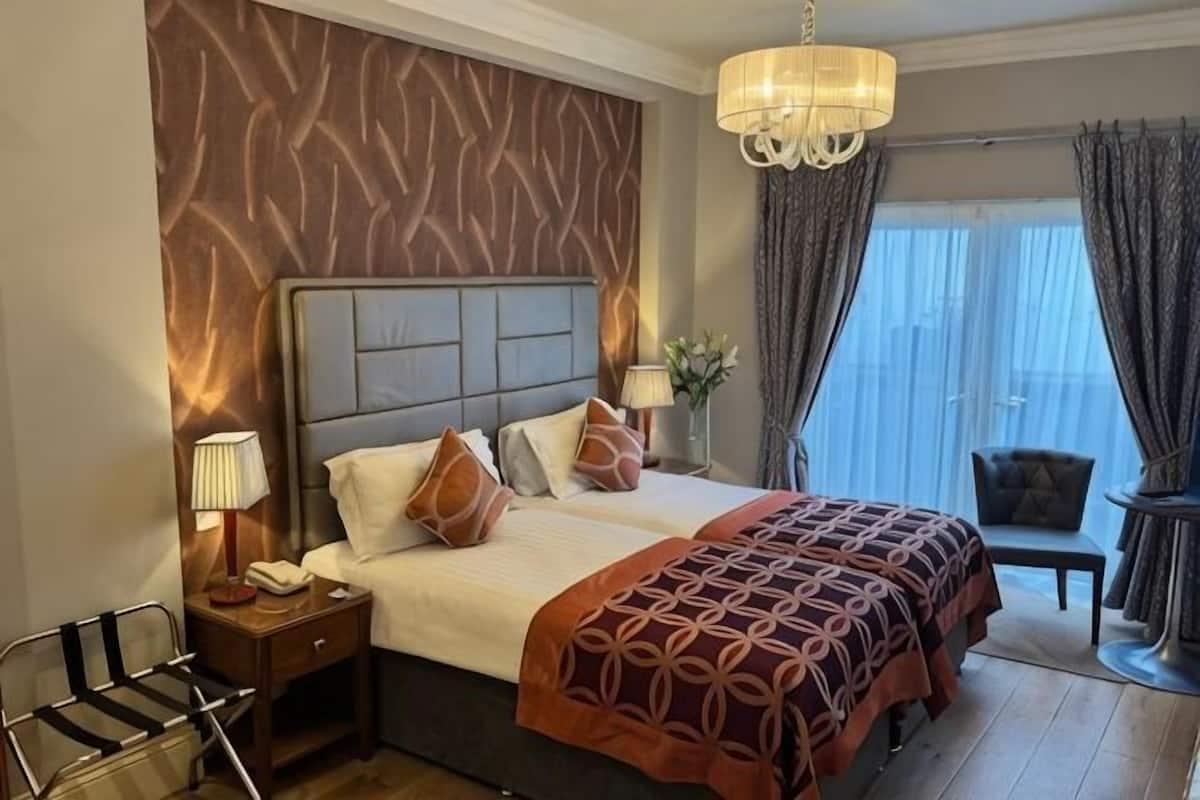 Habitación de hotel con una cama grande, una mesita de noche con lámpara, una silla y vistas al exterior.