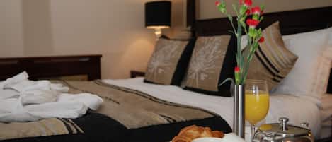 A neatly made bed with a bedside table holding croissants, a cup of coffee, a glass of orange juice, and a vase with flowers.