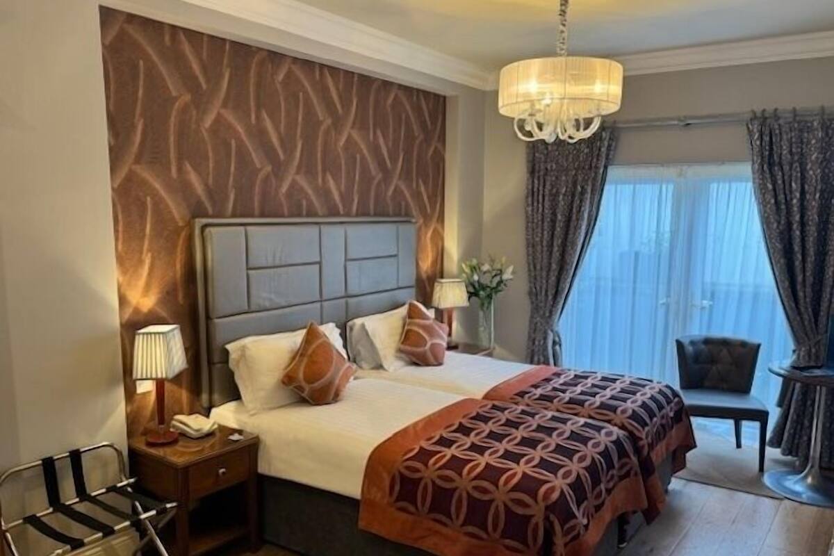 Habitación de hotel con una cama grande, una mesita de noche, una silla, una ventana con cortinas y un candelabro.