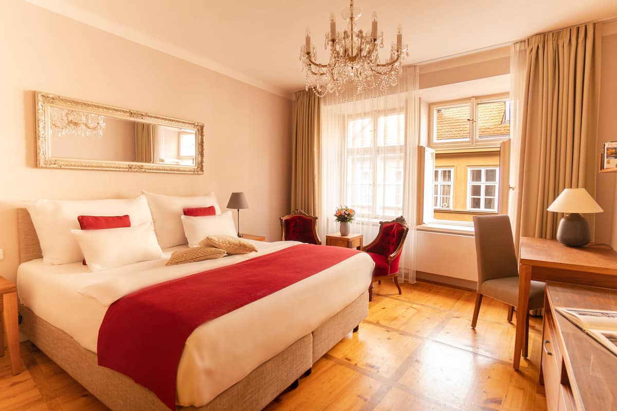 Quarto de hotel com cama grande, lustre, escrivaninha com abajur, cadeira e janela com cortinas.