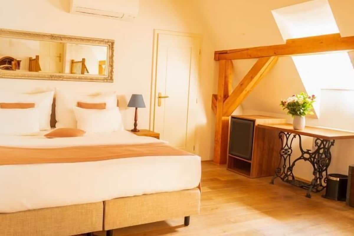 Quarto com cama, duas mesas de cabeceira com luminárias, espelho, escrivaninha de madeira com cadeira e janela com vista.