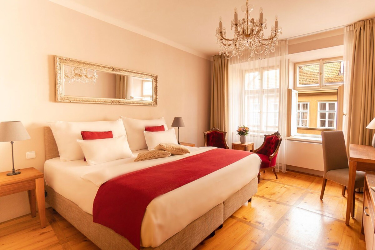 A hotel room with a large bed, a chandelier, a desk with a lamp, a chair, a mirror, and a window with curtains.