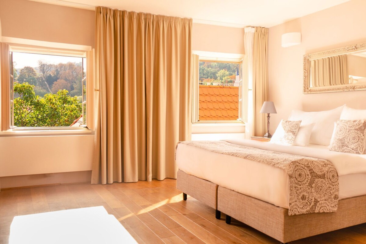 A bedroom with a bed, a nightstand, a lamp, a window with curtains, and a view of greenery outside.