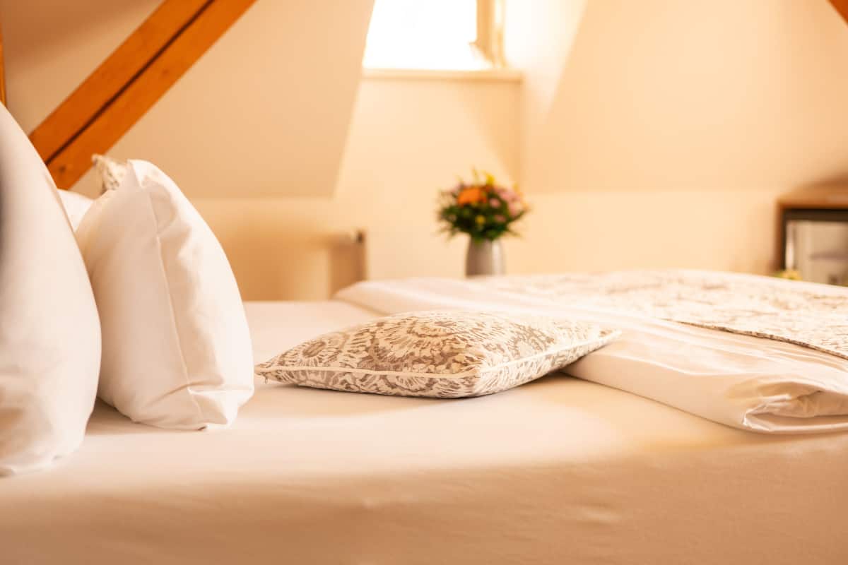 Uma cama bem arrumada com almofadas brancas e estampadas, um cobreleite branco e um pequeno vaso com flores ao fundo.