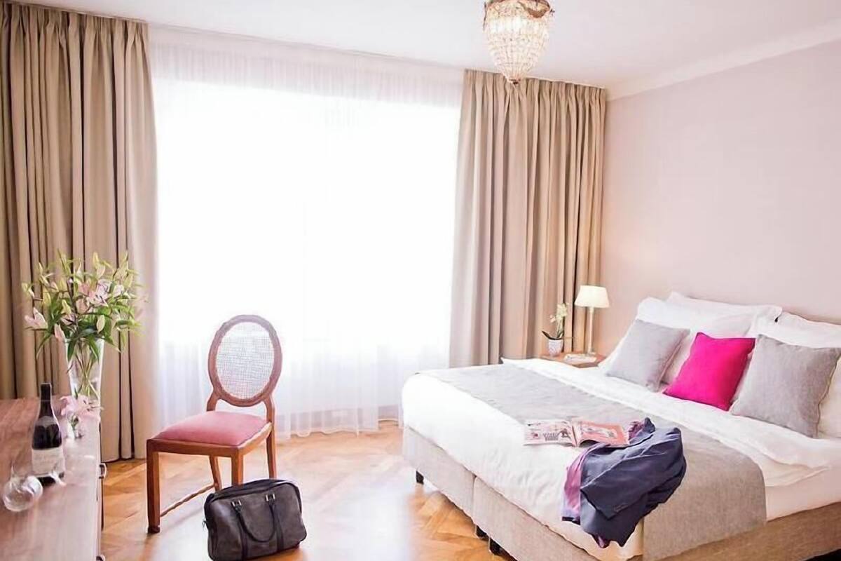 Quarto de hotel com cama, cadeira, escrivaninha, abajur e um vaso com flores.