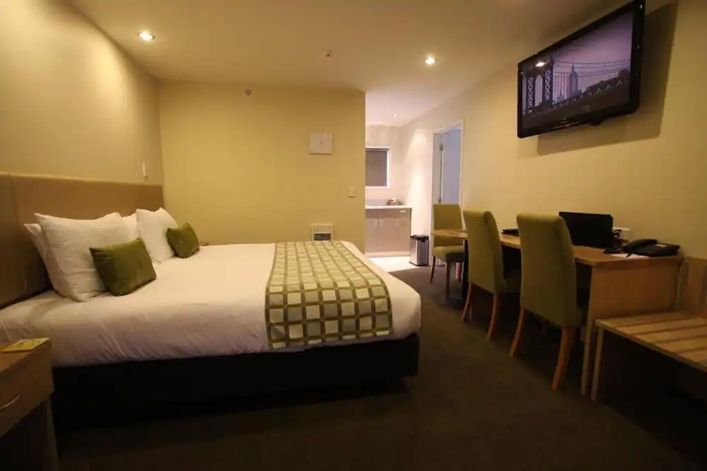 Quarto de hotel com uma cama, uma escrivaninha com cadeira, TV fixada na parede e uma pequena cozinha integrada ao fundo.