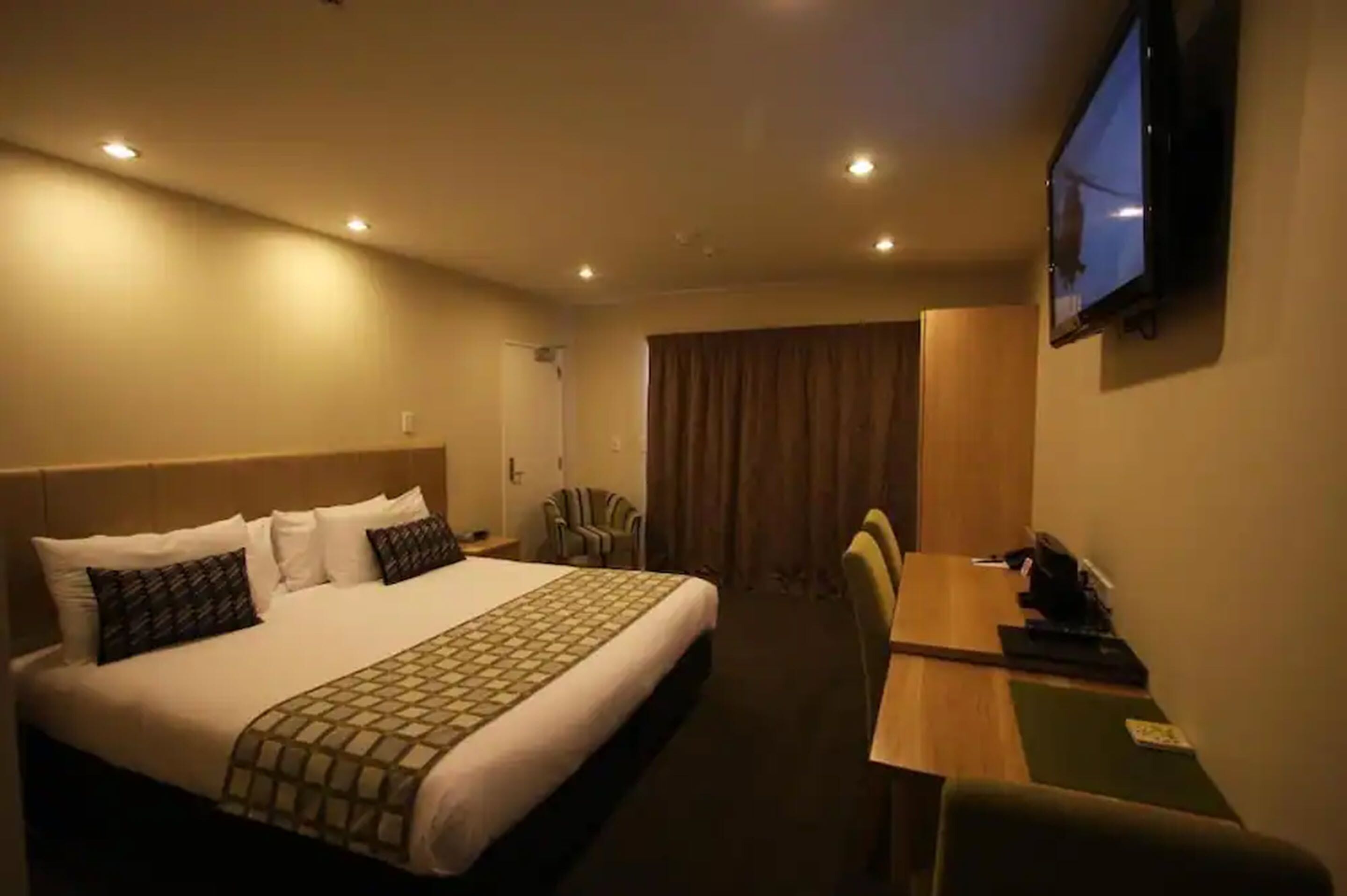 A hotel room with a large bed, a desk, a chair, a TV mounted on the wall, and a window with curtains.