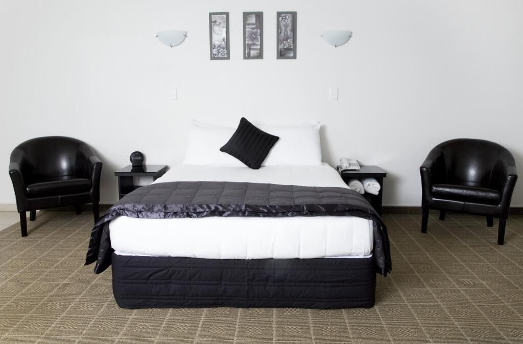 A hotel room with a large bed, two black leather chairs, a nightstand with a lamp, and three framed pictures on the wall.