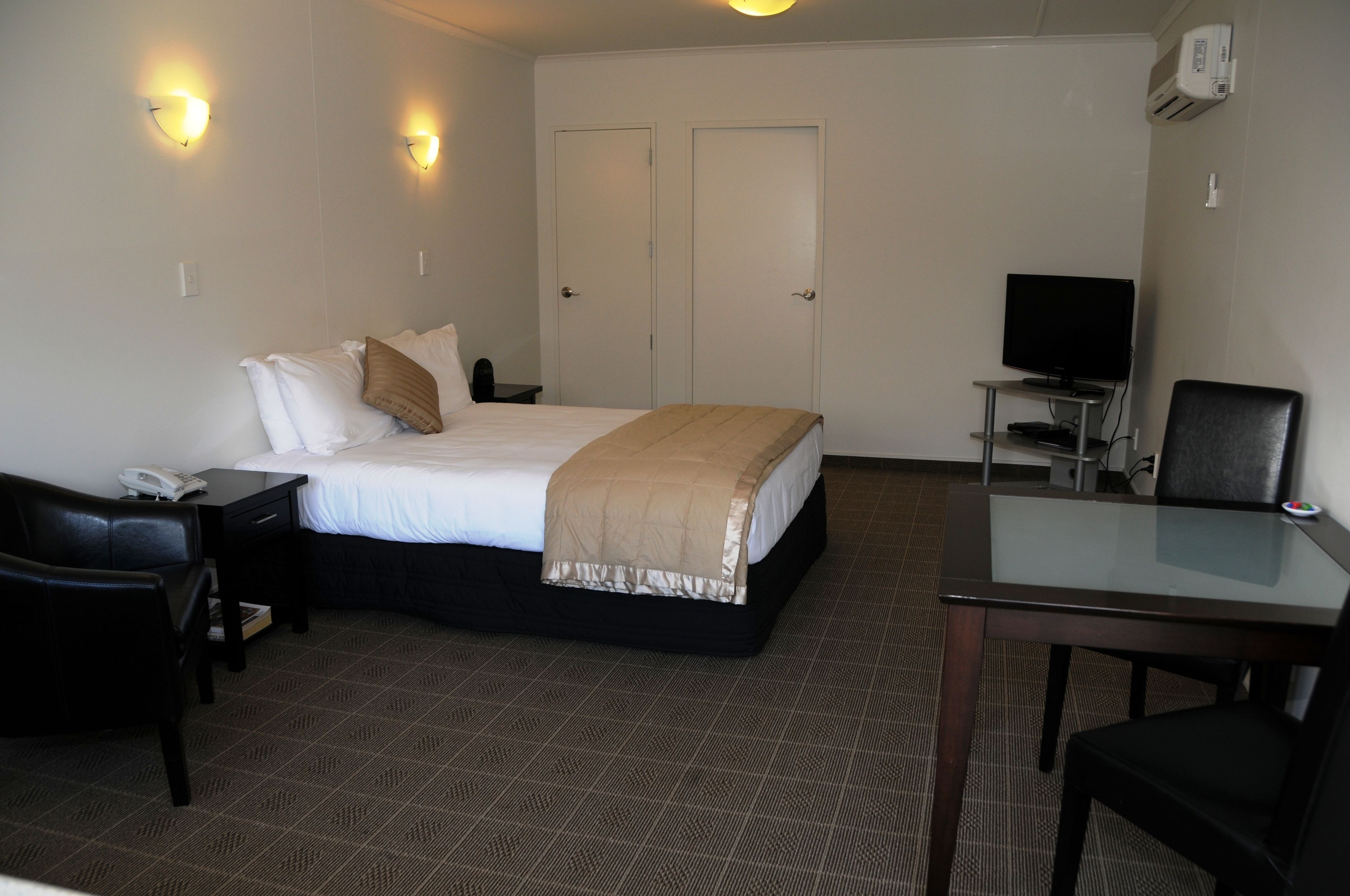 Habitación de hotel con cama, mesita de noche, lámpara, escritorio, silla, televisión y una pequeña mesa con superficie de vidrio.