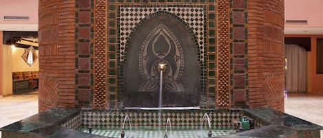 Une fontaine dotée d’une ouverture centrale d’où jaillit l’eau et de plusieurs petites ouvertures supplémentaires, le tout entouré d’une base carrelée ornée d’un motif géométrique.
