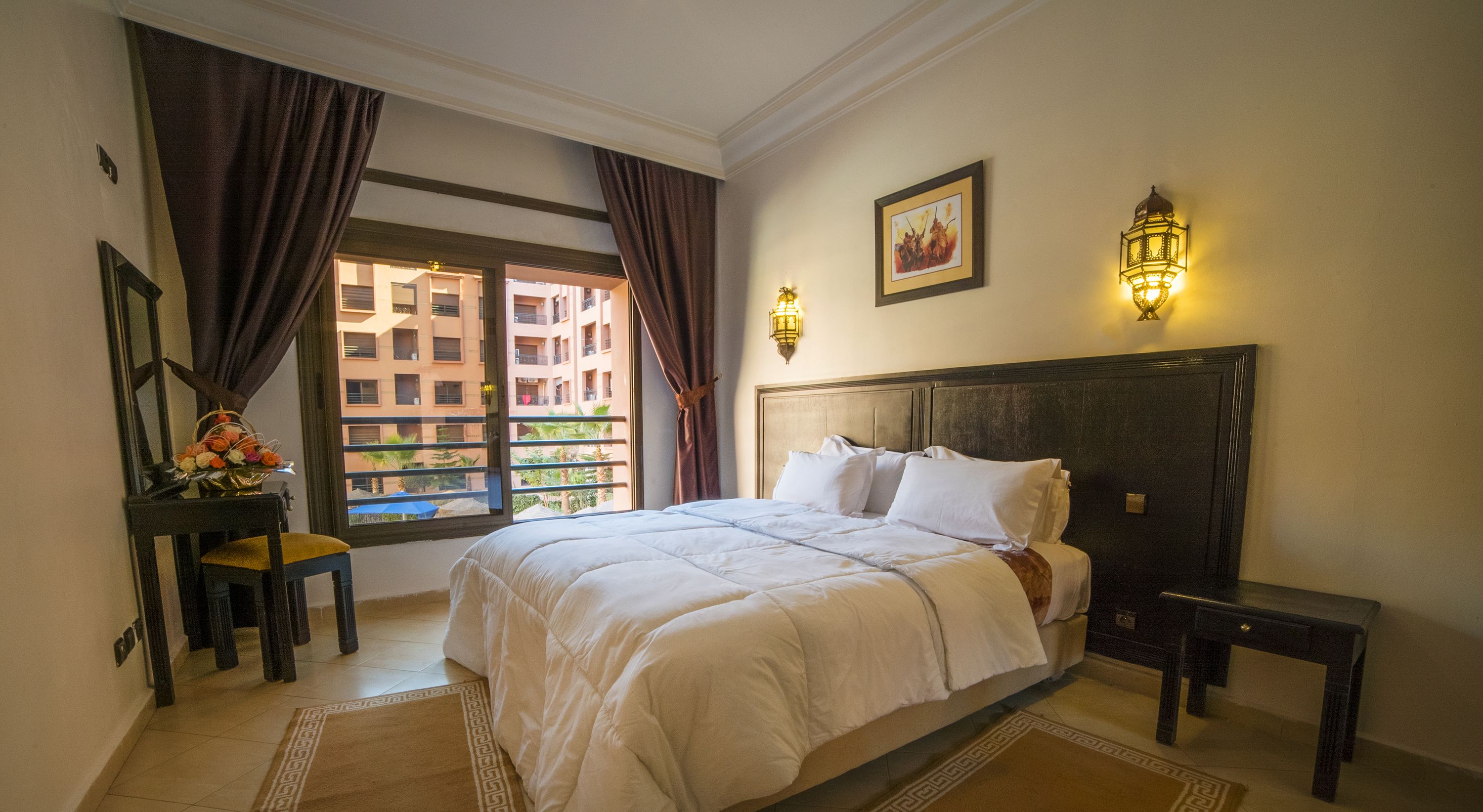 A hotel room with a large bed, a desk with a chair, a window with curtains, and a wall-mounted light fixture.