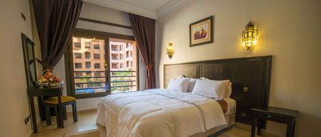 A hotel room with a large bed, a desk with a chair, a window with curtains, and a wall-mounted light fixture.