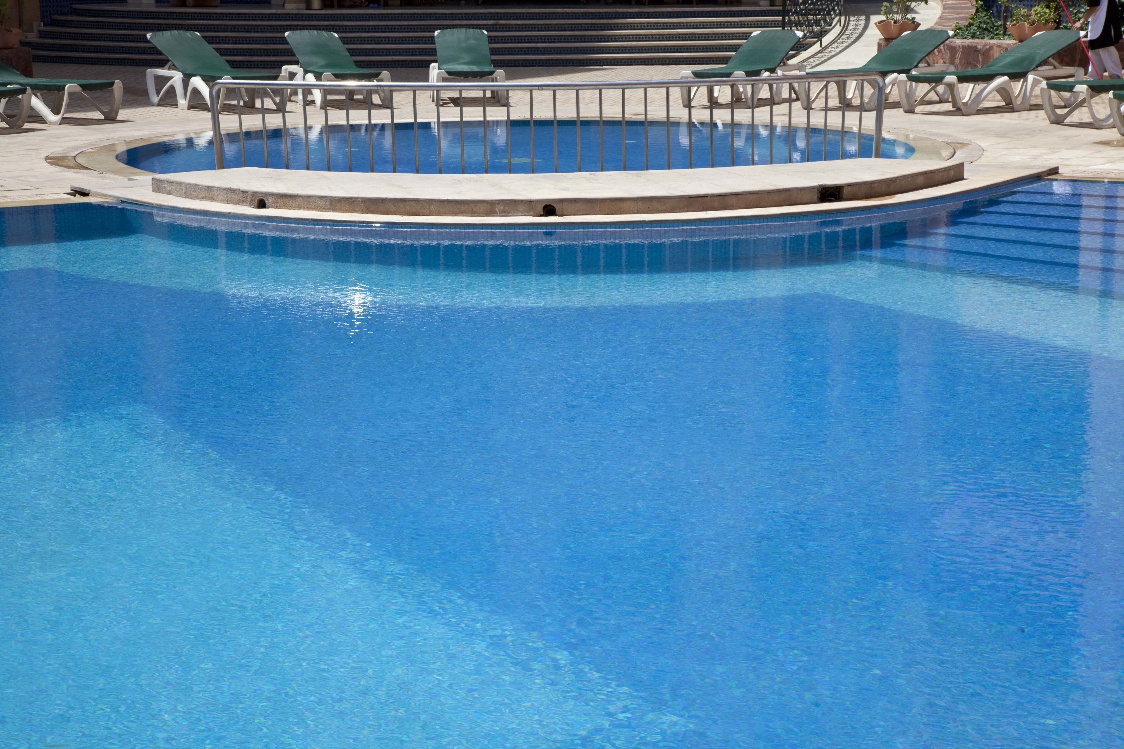 A swimming pool with a circular hot tub in the center, surrounded by green lounge chairs.