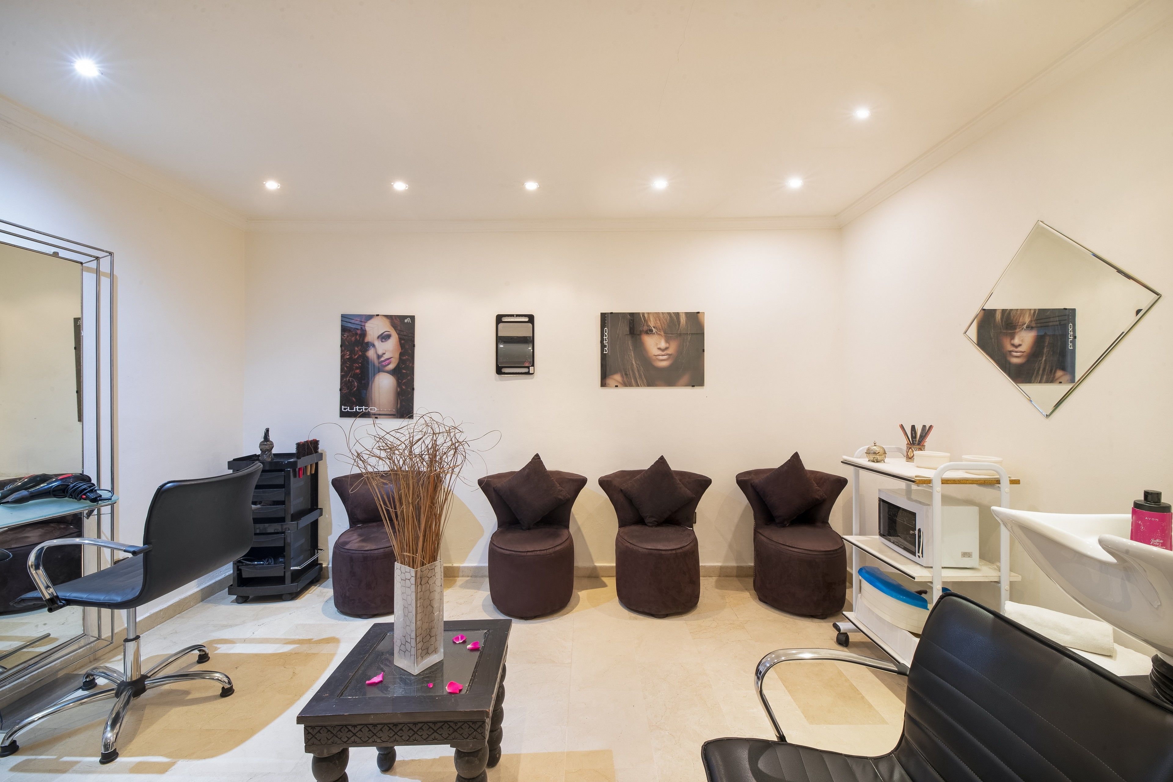A modern salon with a central reception desk, multiple seating areas, and a large mirror on the wall.