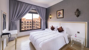 A hotel room with a large bed, a bedside table, a nightstand, a window with curtains, and a view of buildings outside. - Mogador MENZAH (Marrakech)