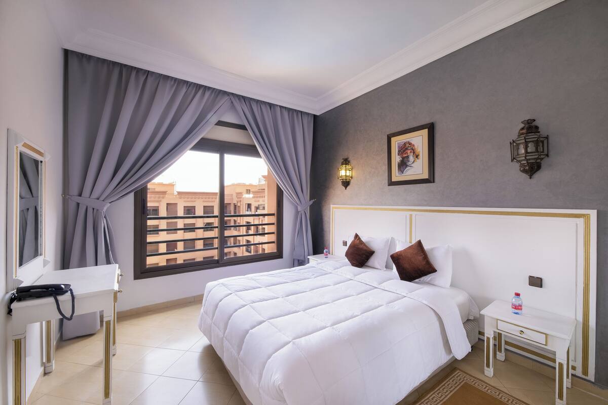 A hotel room with a large bed, a bedside table, a nightstand, a window with curtains, and a view of buildings outside.