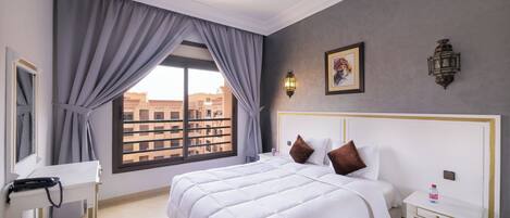 A hotel room with a large bed, a bedside table, a nightstand, a window with curtains, and a view of buildings outside.