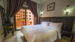 A hotel room with a large bed, a desk with a chair, a balcony with a view of a building, and a wall-mounted light fixture. - Mogador MENZAH (Marrakech)