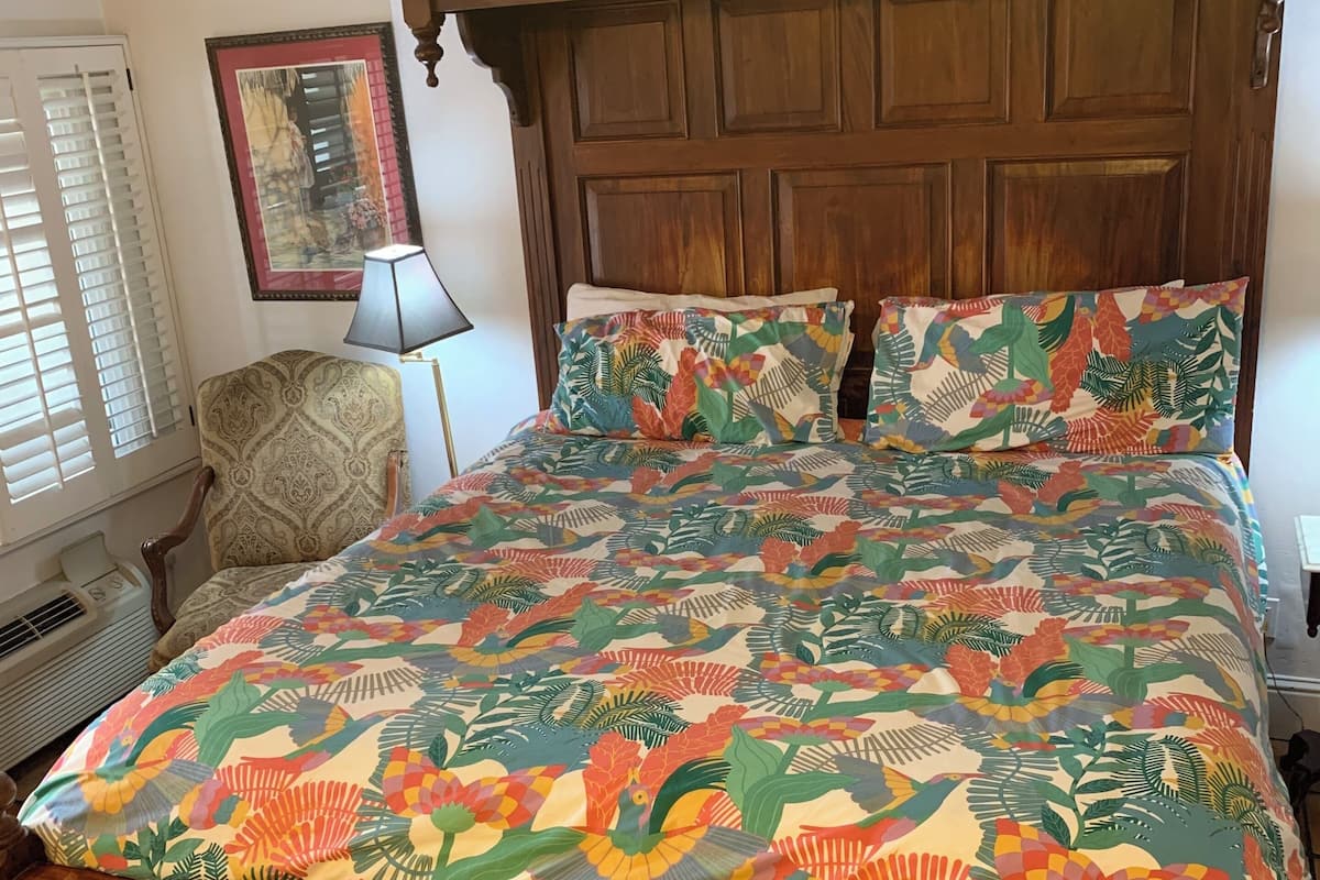 A wooden bed with a floral bedspread and matching pillows, a nightstand, a chair, and a wall-mounted lamp.