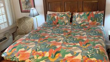 Una cama de madera con colcha y almohadas estampadas, una mesita de noche, una silla y una lámpara de pared.