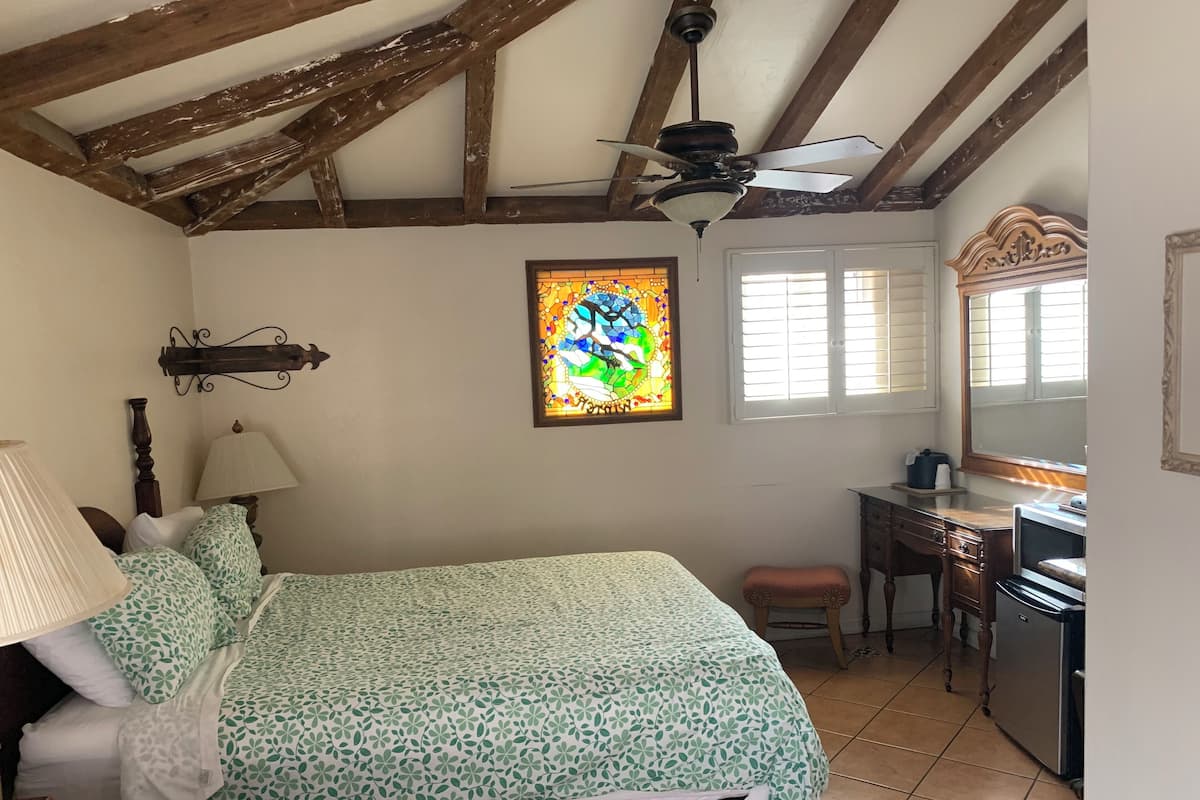 A bedroom with a bed, a nightstand, a lamp, a dresser, a mirror, a window with blinds, a ceiling fan, and a stained glass window.